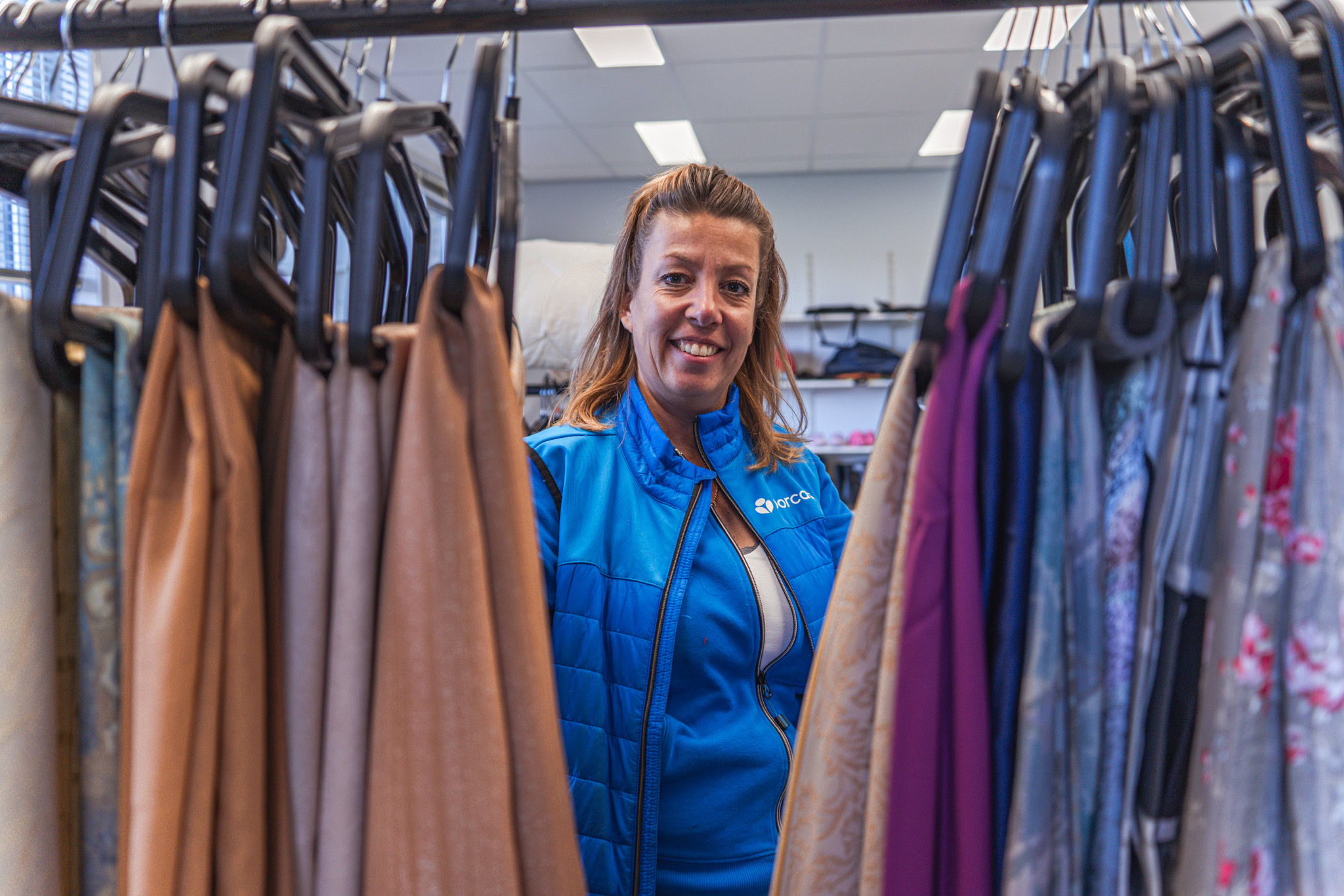 Mkb-merkbare kleding aan haken in kringloopwinkel met lachende medewerker in blauwe jas van Dorcas.