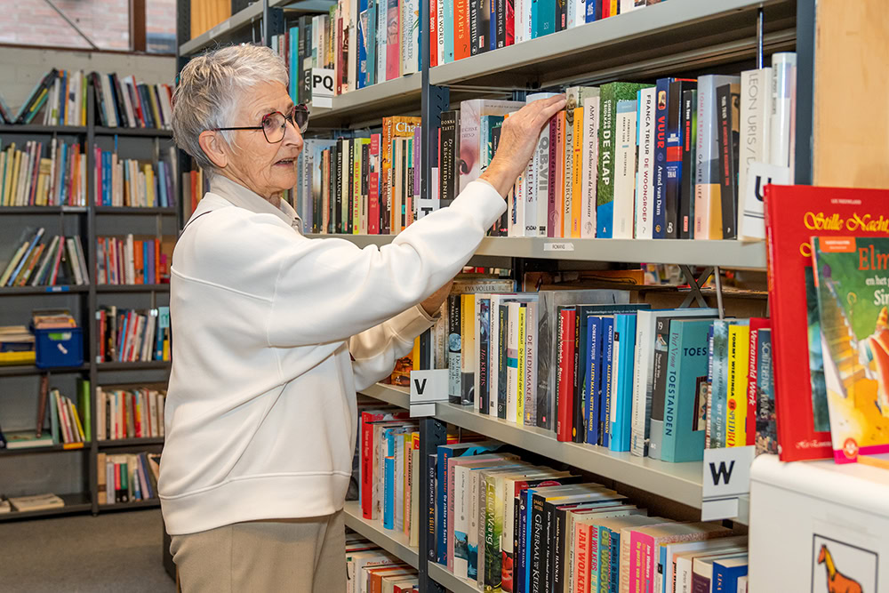 Oudere vrouw zoekt een boek in een bibliotheek, omringd door kleurrijke boekenkasten met diverse boeken.