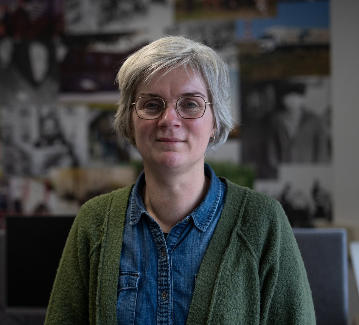 Inspirerende vrouw met korte blonde haar en brillen, zittend in creatieve kantooromgeving met posters op de achtergrond, representatief voor maatschappelijke betrokkenheid bij Dorcas.