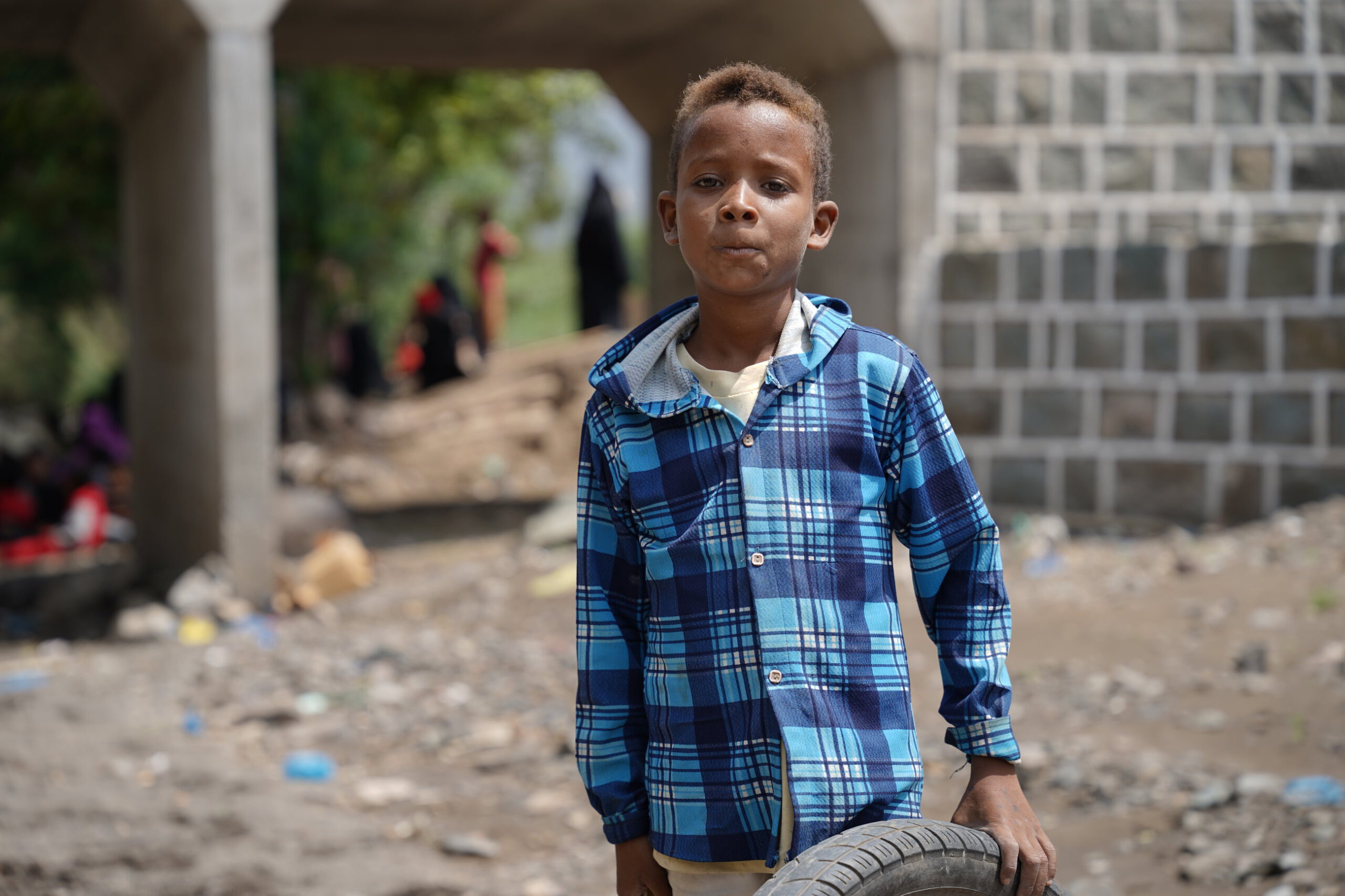 Kind met een blauwe geruite jas die een autoband vasthoudt naast een brug.