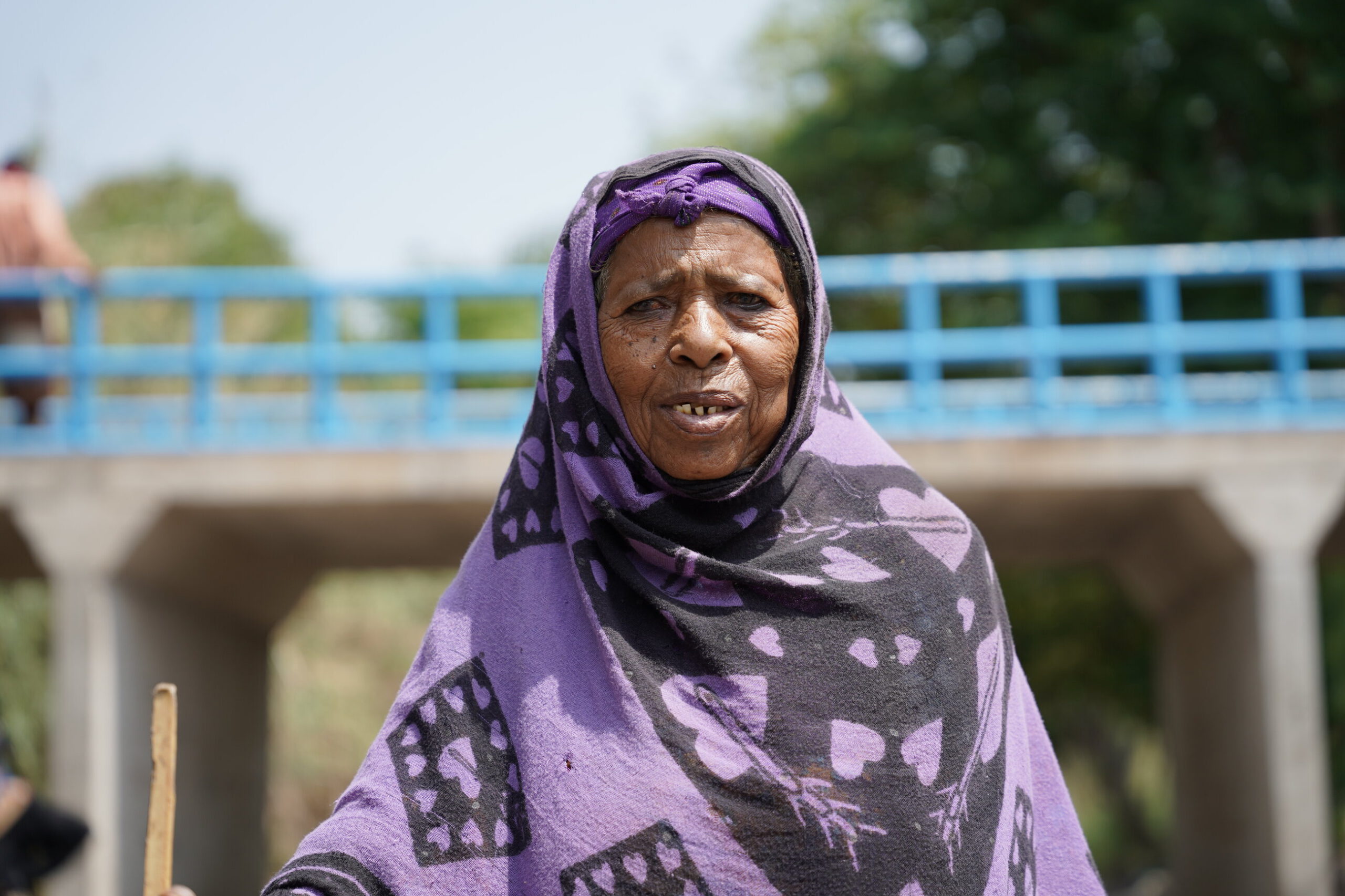 Levende portret van een oudere vrouw in kleurrijke hoofddoek, naast een brug