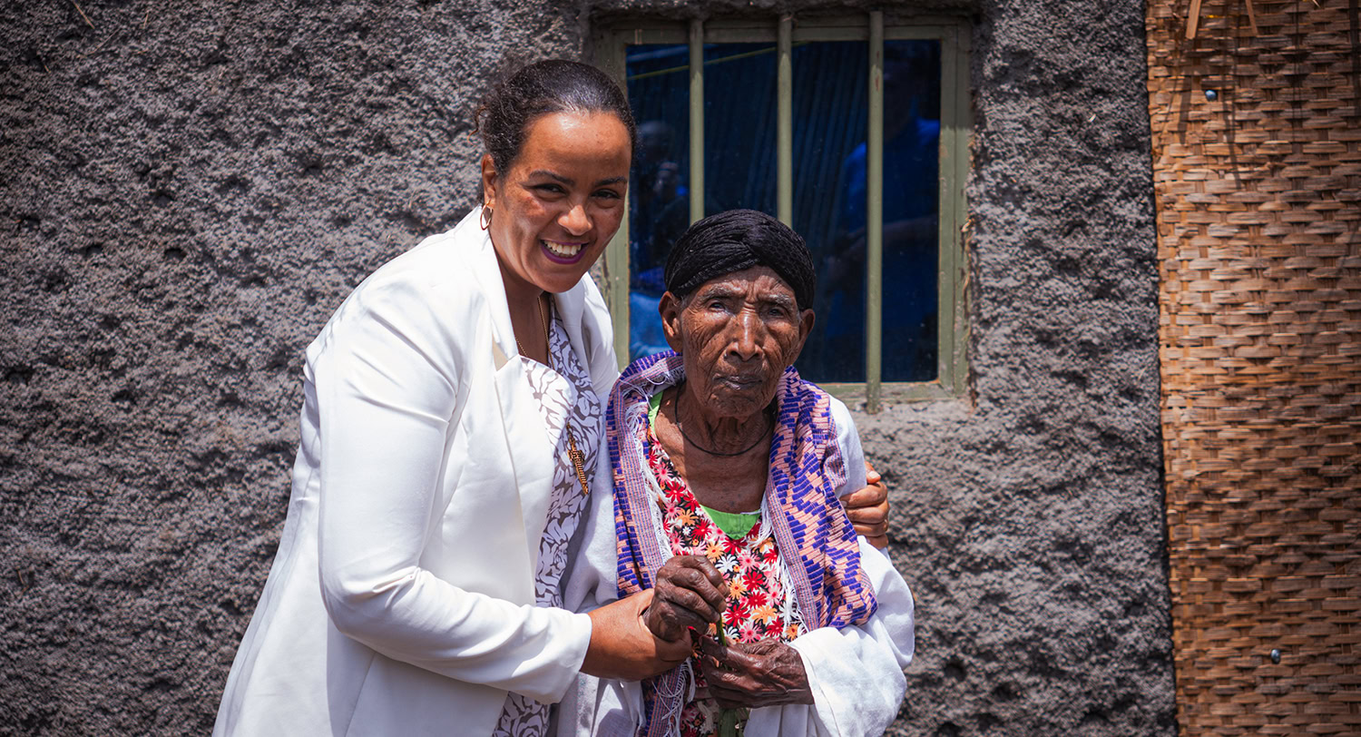 Vrouwelijke hulpverlener ondersteunt oudere vrouw in rural gebied in Kenia, verantwoordelijk voor lokale sociale en medische zorg, dorpshuis in de achtergrond.