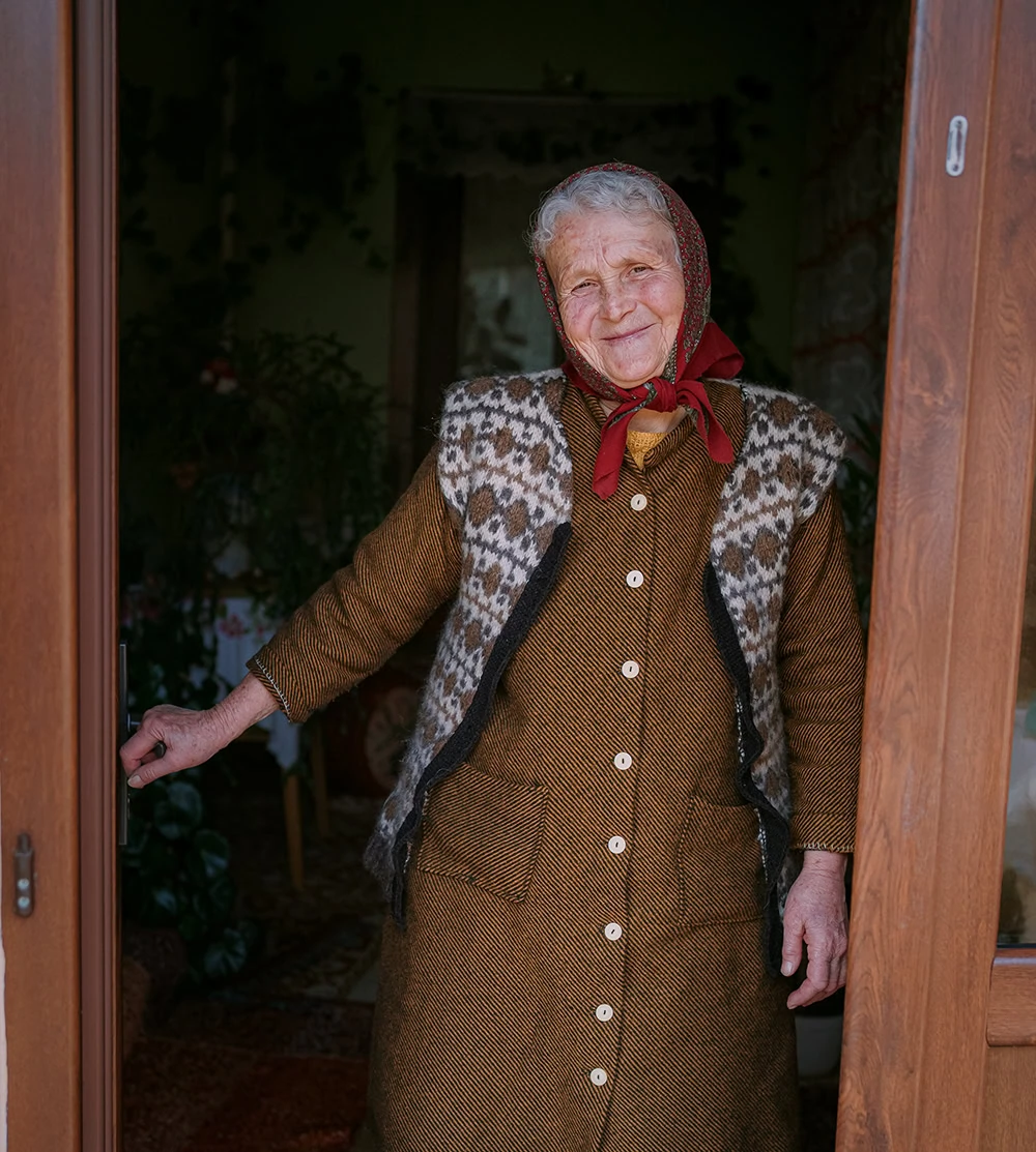 Oudere vrouw met warme kleding die vriendelijk lacht, bij de ingang van haar huis.