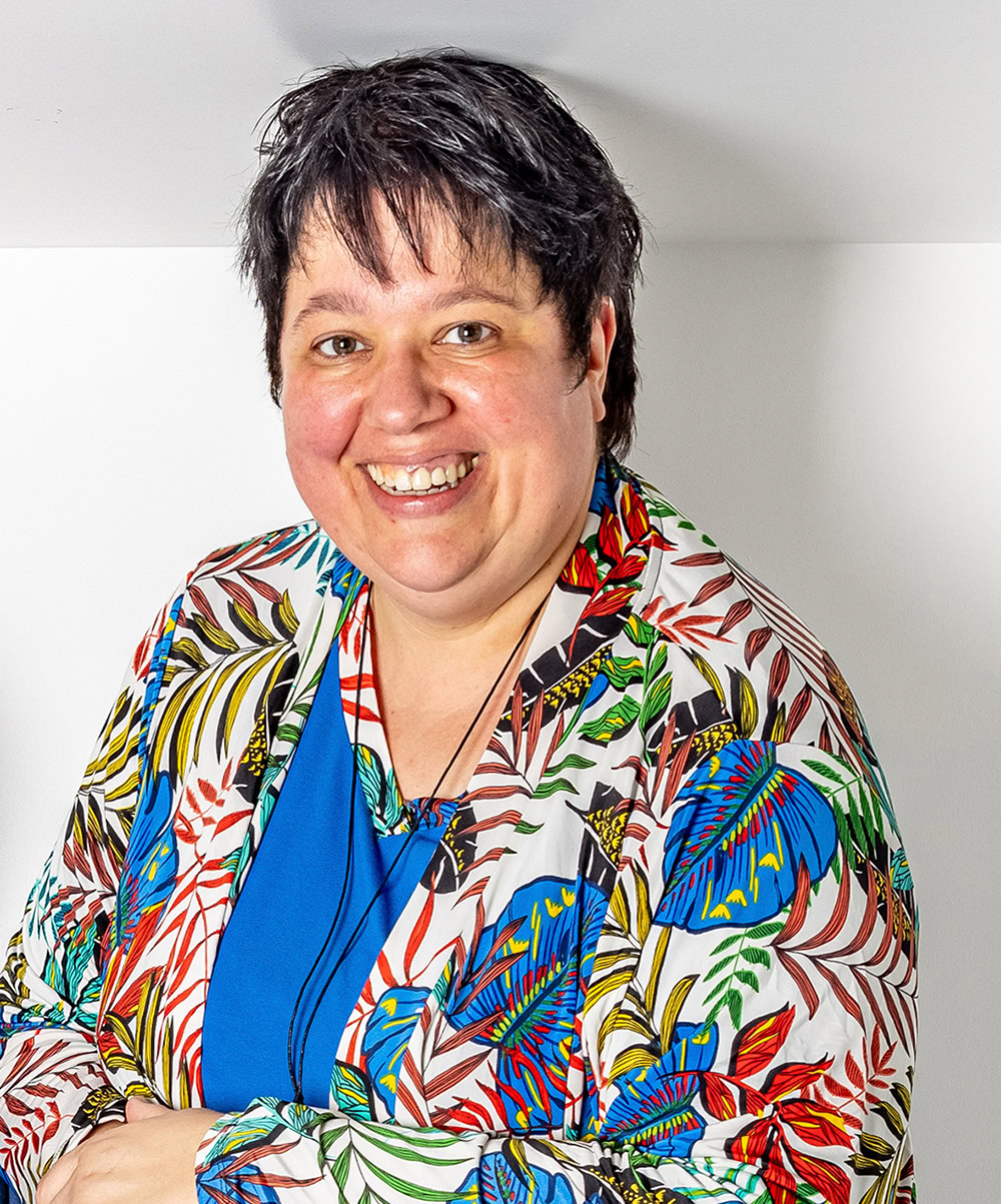 Blije vrouw met kort zwart haar draagt kleurrijk jungleprint jasje en blauwe blouse, vrolijk glimlachend, portret op lichte achtergrond, mode en geluk.