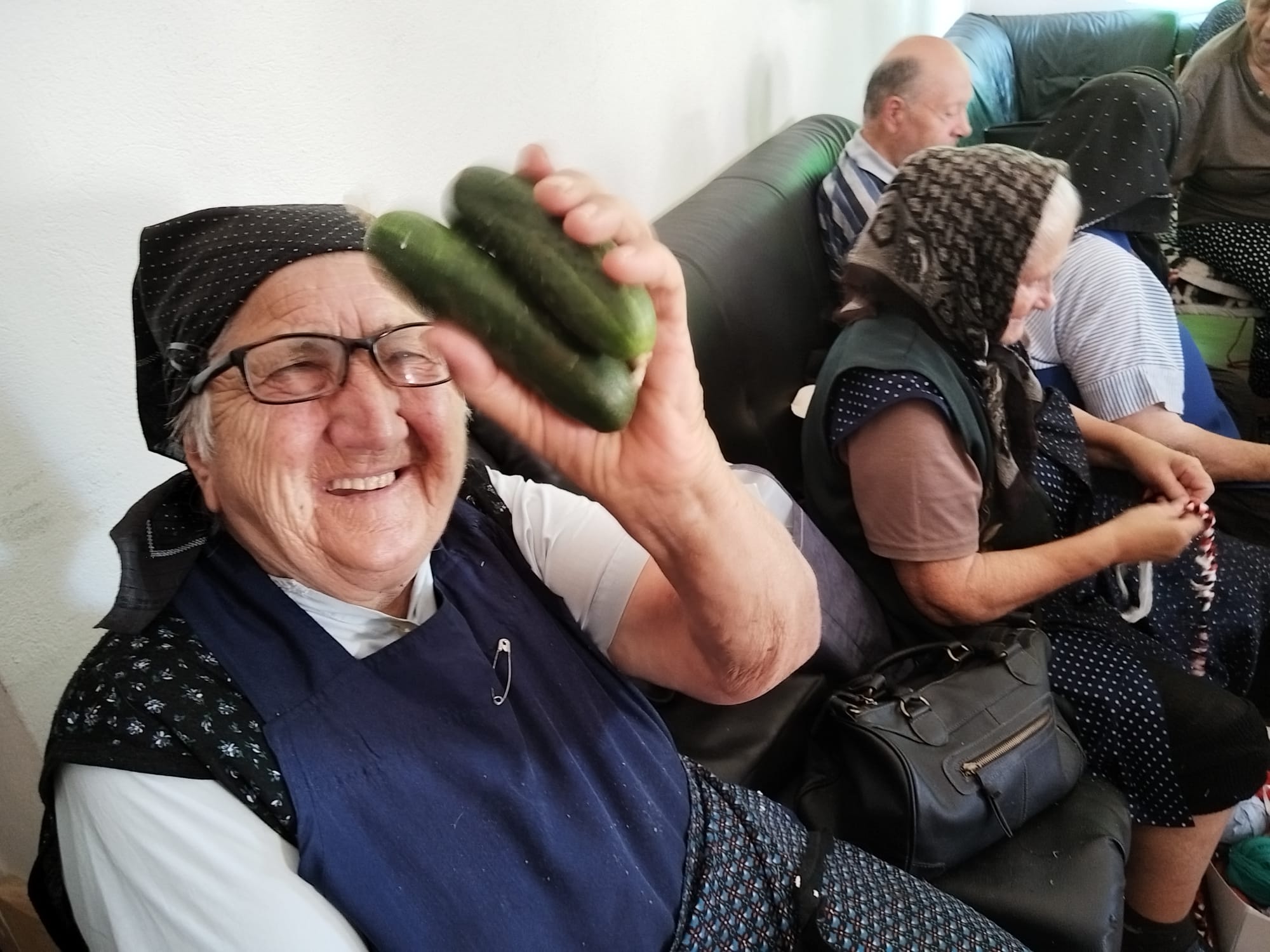 Vrolijke oudere vrouw met brillen en kopdoek die komkommers vasthoudt op een gezellige groep van ouderen op een bank.