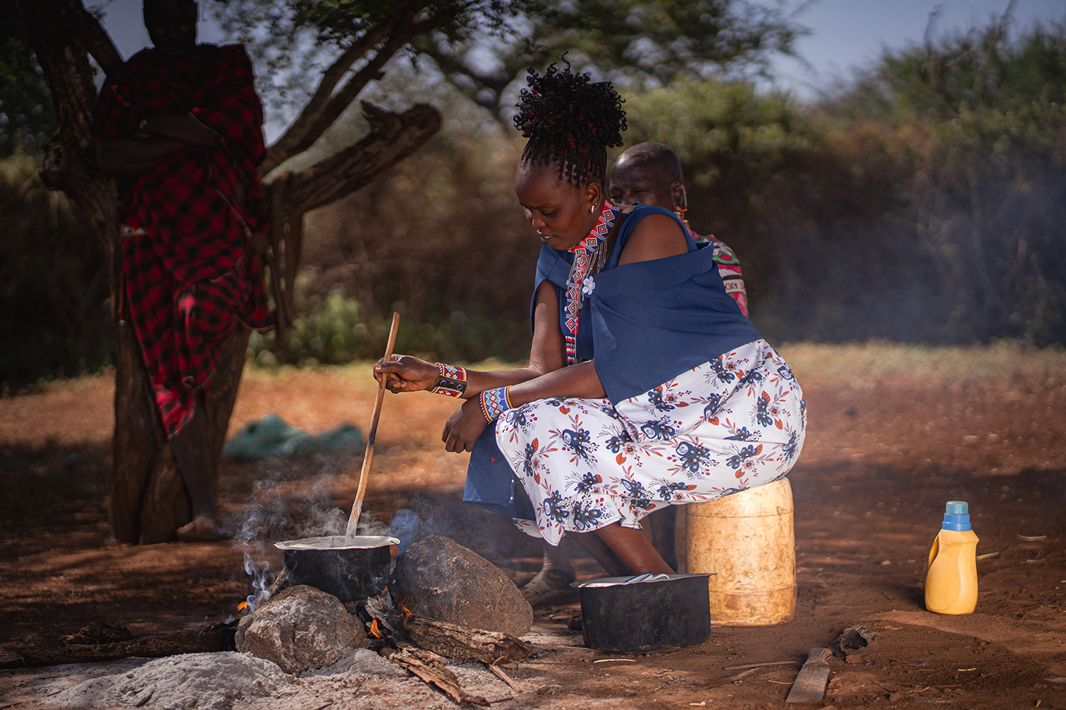Grote kleurrijke kralen armband en sjaal, vrouw kookt op open vuur in een buitenomgeving, traditionele Afrikaanse kleding en accessoires, verbonden met dorcas.nl voor ontwikkelingshulp en verbetervoorbeelden.