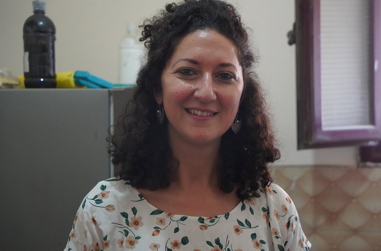 Vrouw met donkere, krullende haar lachend in een keukenruimte, draagt een bloemenprint blouse, symboliseert betrokkenheid en solidariteit bij projecten van Dorcas.